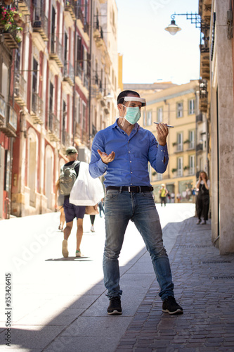Uomo con mascherina facciale e visiera facciale in plexiglass parla al cellulare mandando un messaggio vocale in centro città
