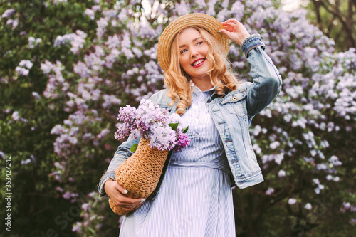 Portrait young happy attractive blonde woman in denim jacket, dress posing with purple flowers in straw bag in countryside park. Beautiful cheerful hipster girl have fun, relax on blossom lilac garden