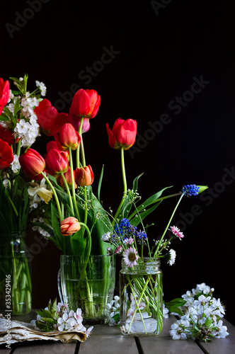 Still life with bouquets of red tulips, field daisies, muscaris in glass jars, cherry blossoms on dark background