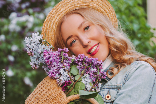Portrait beautiful young happy blonde woman with wavy hair posing with bouquet of lilac flower in straw bag on blossom garden. Cheerful carefree pretty hipster girl relax, enjoy floral nature outdoors
