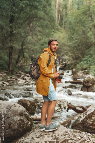 Tourist man photographer wearing a yellow coat with camera in the middle of the forest with moutain river. Beautiful summer colors.
