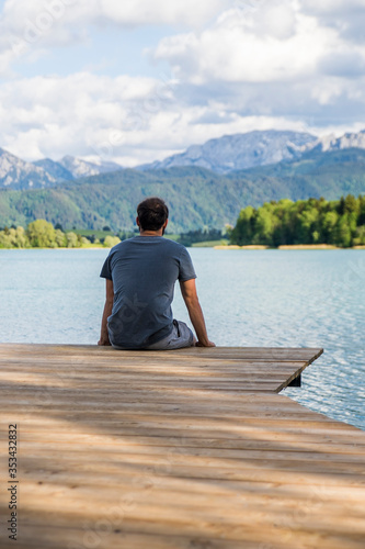 Mann sitzt auf einem Bootssteg mit einer schönen Alpenlandschaft und einem See