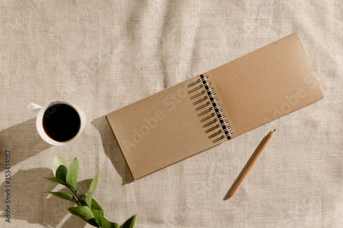 Blank brown vintage sketchbook, pencil, cup of coffee, green leaves on beige linen fabric in the morning. Minimalistic workplace concept. Top view on background with copy space. Summer flat lay