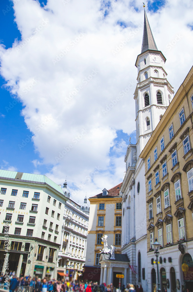 Naklejka premium Old city scape with church tower on right side and urban architecture of Vienna city, Austria. History concept with tourists on the street. Blue sky and white clouds above the street.