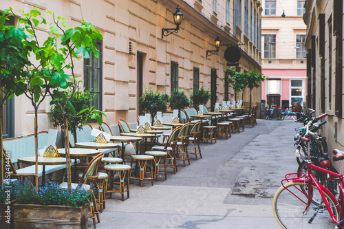 Green chairs with brown chairs on the small street, on left side are standing bicycles. Touristic road in Vienna, Austria. Old narrow street as European cityscape. 
