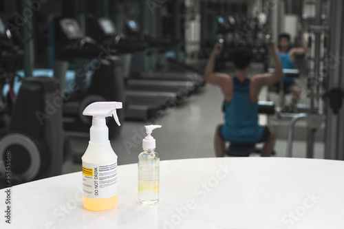 Close up spray bottle of yellow disinfectant and alcohol gel with blurred background of Asian man workout in gym. Concept for prevention and protection of Corona virus Covid 19 in fitness center.
