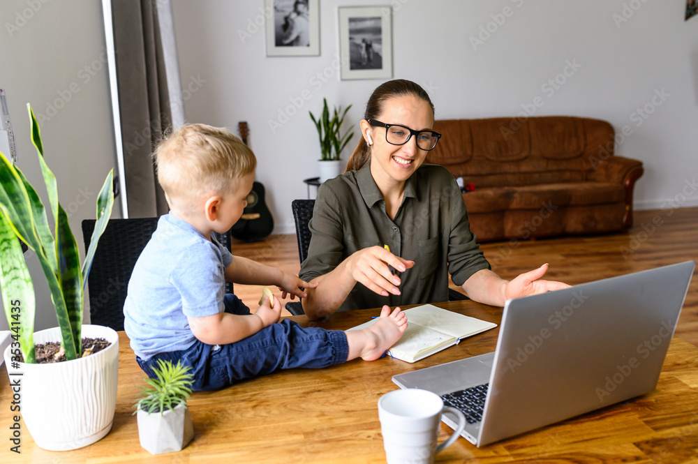 Pretty young mother in eyeglasses is talking online, she using laptop for video meeting, cute baby boy sits on a table near. Busy mom