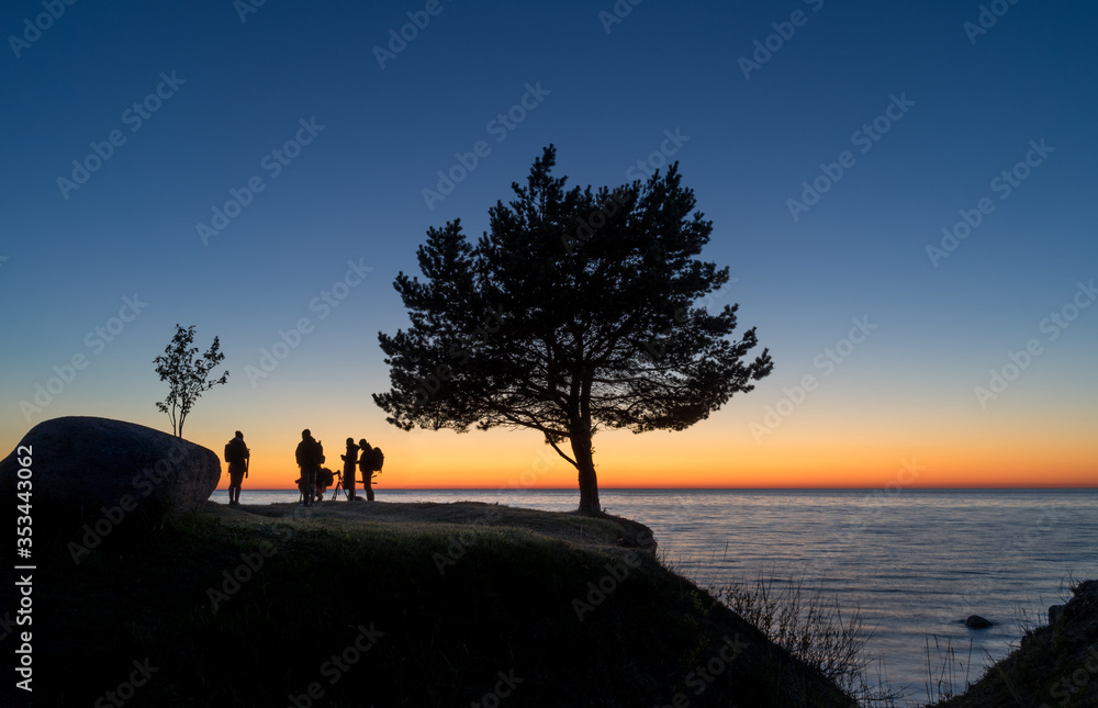 Late evening at the sea. Orange sundown stripe at the horizon. Dark silhouette of single hardwood tree and few people in front of dawning skyline. Calm water of Finnish Gulf. Peaceful twilight sky. Late evening at the sea. Orange sundown stripe at the horizon. Dark silhouette of single hardwood tree and few people in front of dawning skyline. Calm water of Finnish Gulf. Peaceful twilight sky.