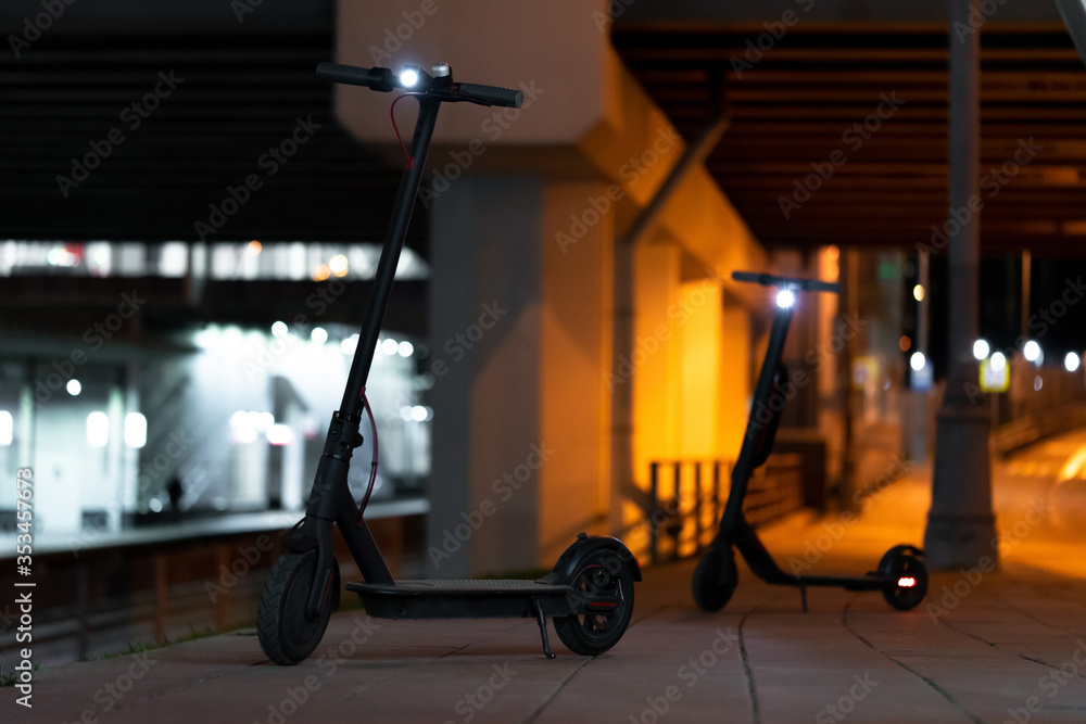 Black Electric Kick Scooters With Turn On Headlamps at Cityscape at Night Time. Scooters Under Illuminated Bridge And Near Metro Station. Urban Transportation