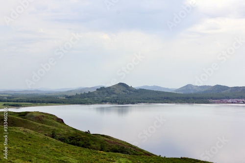 Panoramic view. A lagoon with a clean mountain lake in the midst of majestic mountains in a haze of fog. Green grass. Apartments around the lake