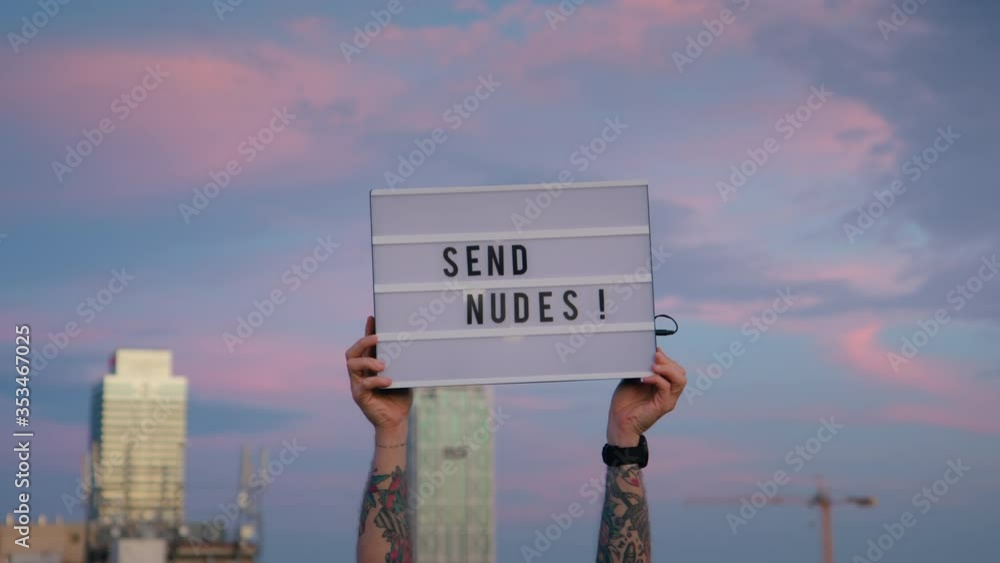 Funny sign in sky say send nudes, modern memes concept. Man holds up in city sunset sky lit up
