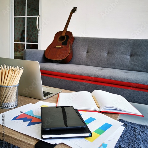 notebook computer with pencils and mobile telephone on the working table with guitar on the couch in living room. work from home concept