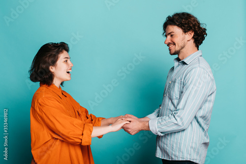 Side view of young couple standing together and look at each other. Holding hands and smiling. Proposal time in studio. Isolated over blue background.