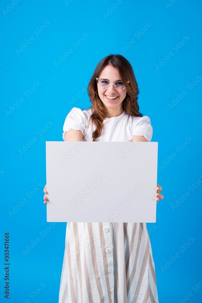 Happy young brunette woman girl in summer clothes toothy smile isolated on blue background studio portrait.Holding white big sign board blank paper overhead. Dentistry, shopping discount sale Happy young brunette woman girl in summer clothes toothy smile isolated on blue background studio portrait.Holding white big sign board blank paper overhead. Dentistry, shopping discount sale