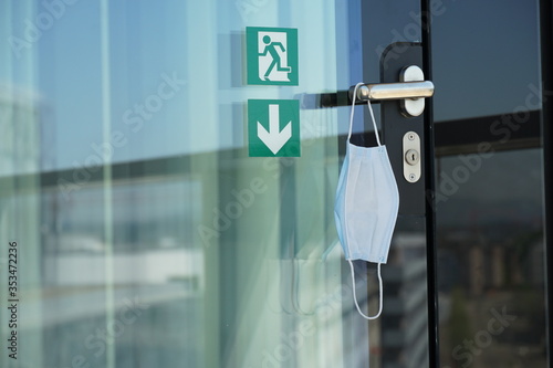 A door with sign exit with a face mask suspended on a door handle in a corporate building, close up. A reminder of safety measures in prevention of coronavirus COVID-19 infection. 