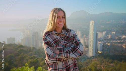 Cute and pretty, young beautiful woman or millennial zoomer teenager stand on top of hill over epic cinematic panorama of big city. Woman laugh and smily shily look at camera on sunny day photo