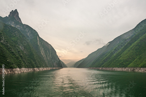 Wushan, Hubei, China - May 7, 2010: Wu Gorge in Yangtze River. Long view with orange lighted sky over green water into the canyon made by green forested mountains with some cliffs.
