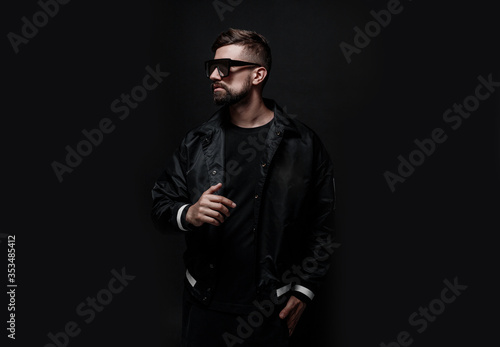 Young man with beard in black jacket and glasses. Portrait of guy on dark background.
