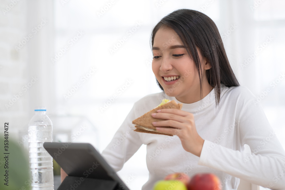 Young Asian woman working with tablet at home and eating sandwich salad fruits at home Young Asian woman working with tablet at home and eating sandwich salad fruits at home