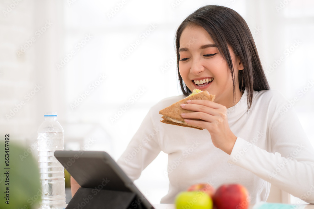 Young Asian woman working with tablet at home and eating sandwich salad fruits at home Young Asian woman working with tablet at home and eating sandwich salad fruits at home