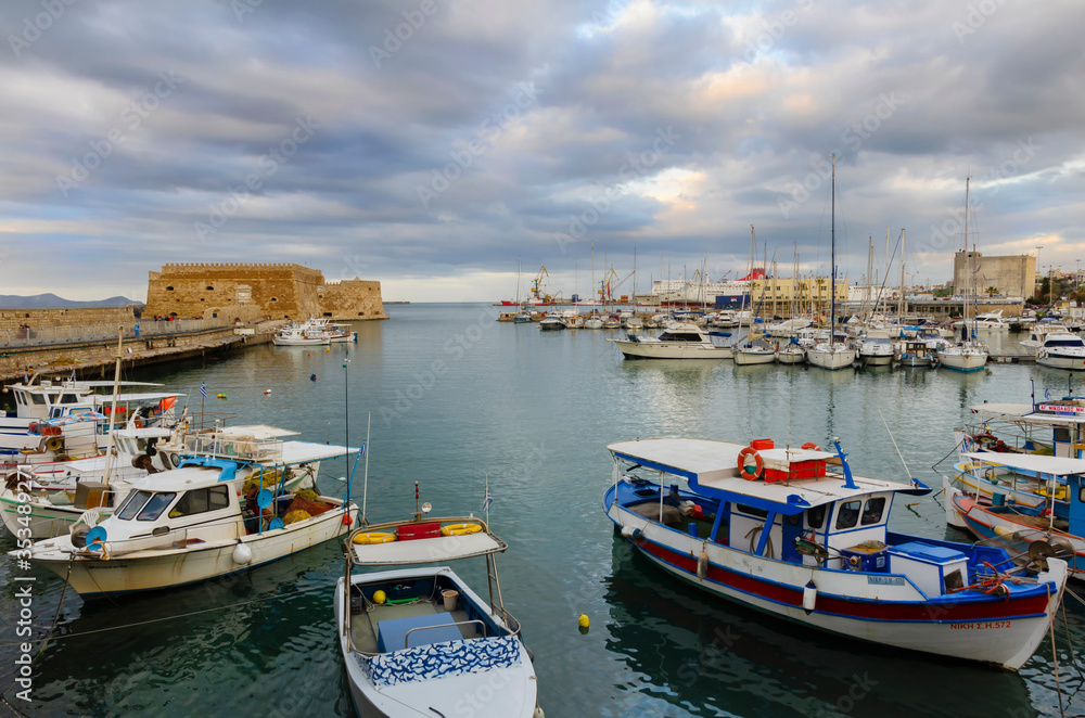 Heraklion, Crete / Greece. Panoramic view at the old Venetian port. Traditional and colorful fishing boats are docked. Venetian fortress Koules in the background. Sunset with cloudy sky