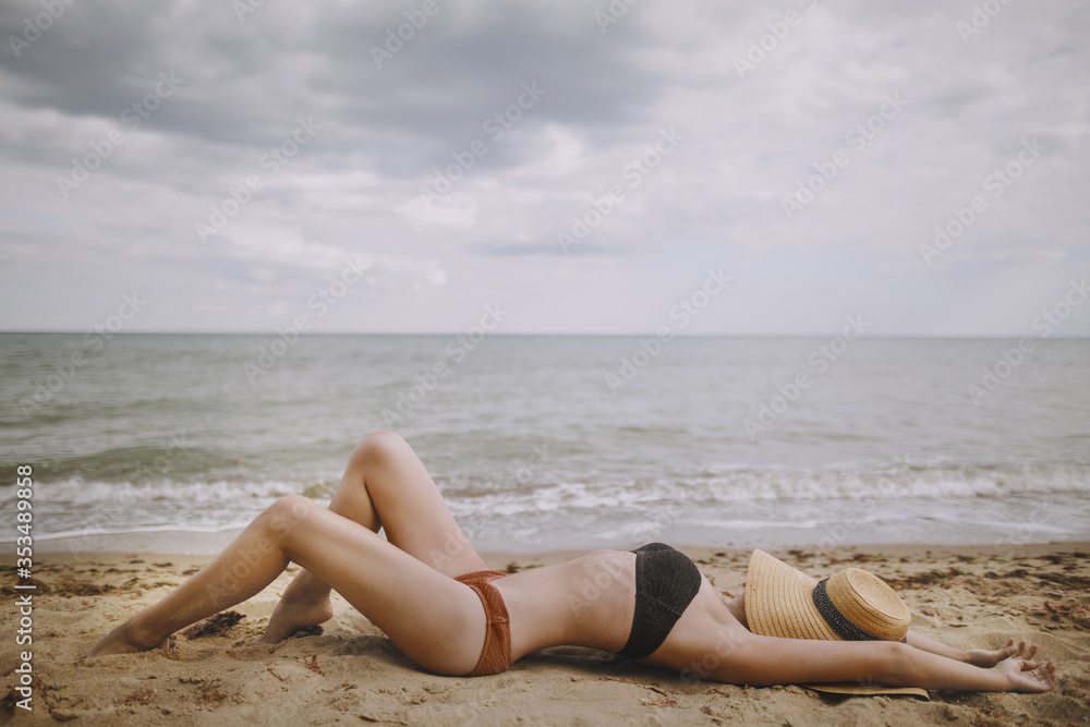Tanned girl in hat lying on beach. Fashionable young woman covering with straw hat, relaxing on sandy beach near sea. Summer vacation and travel. Mindfulness and carefree Tanned girl in hat lying on beach. Fashionable young woman covering with straw hat, relaxing on sandy beach near sea. Summer vacation and travel. Mindfulness and carefree