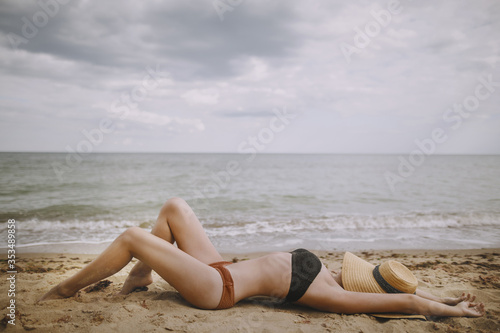 Tanned girl in hat lying on beach. Fashionable young woman covering with straw hat, relaxing on sandy beach near sea. Summer vacation and travel. Mindfulness and carefree