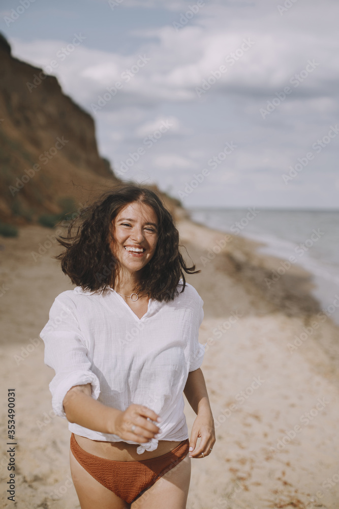 Joyful boho girl in white shirt running on sunny beach. Carefree stylish woman in swimsuit and shirt relaxing on seashore. Summer vacation. Lifestyle authentic image Joyful boho girl in white shirt running on sunny beach. Carefree stylish woman in swimsuit and shirt relaxing on seashore. Summer vacation. Lifestyle authentic image