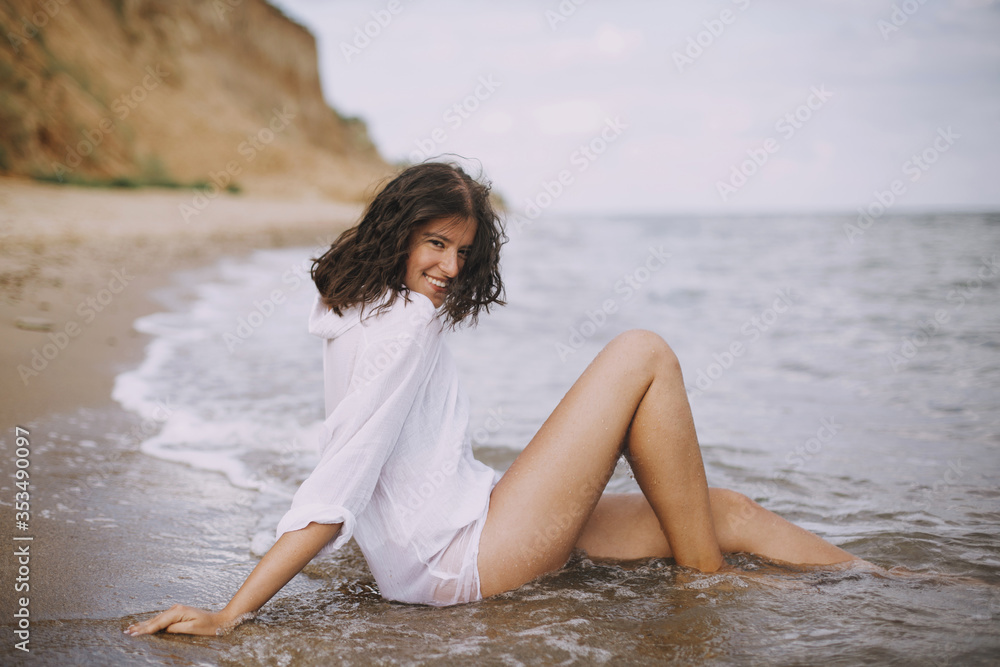 Happy young woman in white shirt sitting on beach in splashing waves. Stylish tanned girl relaxing on seashore and enjoying waves. Summer vacation. Mindfulness and carefree moment Happy young woman in white shirt sitting on beach in splashing waves. Stylish tanned girl relaxing on seashore and enjoying waves. Summer vacation. Mindfulness and carefree moment