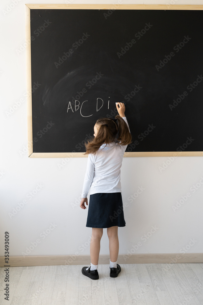 Clever girl in the school office. She stands in front of the board with her back to the camera. The schoolgirl has a uniform, and on her head hair is piled up in tails. Writes English letters. Studyin Clever girl in the school office. She stands in front of the board with her back to the camera. The schoolgirl has a uniform, and on her head hair is piled up in tails. Writes English letters. Studyin