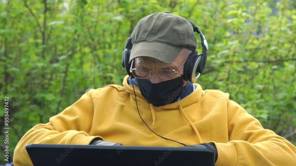 pensioner freelancer in yellow hoodie cap mask gloves and headphones works on notebook in garden at self-isolation closeup pensioner freelancer in yellow hoodie cap mask gloves and headphones works on notebook in garden at self-isolation closeup
