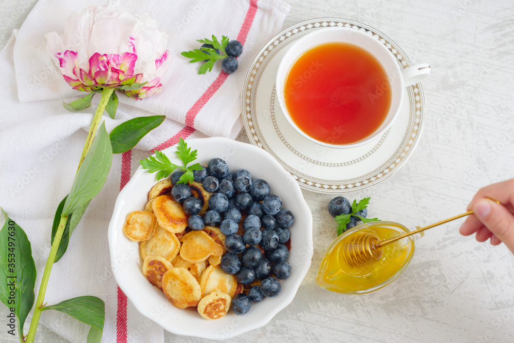 Tiny pancake cereal with blueberry, honey and tea for breakfast in the morning light and pink peony on table Tiny pancake cereal with blueberry, honey and tea for breakfast in the morning light and pink peony on table