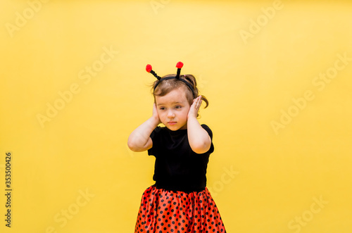 A beautiful girl in a ladybug costume covered her ears against a yellow isolated background with space for text.