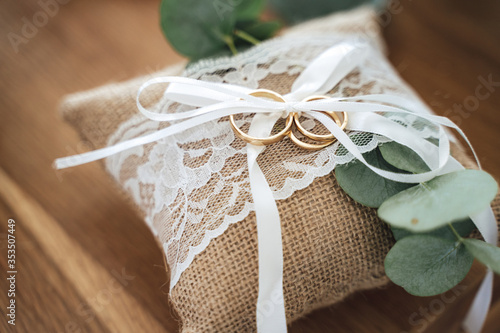 Golden three wedding rings set on small burlap pillow decorated with white lace and ribbon. Wooden background. Wedding day concept.