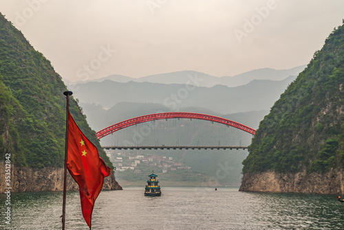Wushan, Chongqing, China - May 7, 2010: Wu Gorge in Yangtze River. Red S103 road bow bridge over Daning River at connection with green Yangtze. Chinese flag and boat. Mountains on horizon. photo
