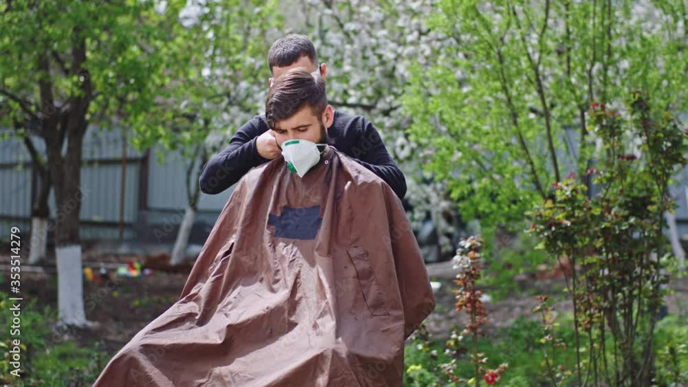 In a large garden two friends handsome guys with a protective mask have a haircut time one of them cut hair to other in quarantine at home in the garden Covid-19