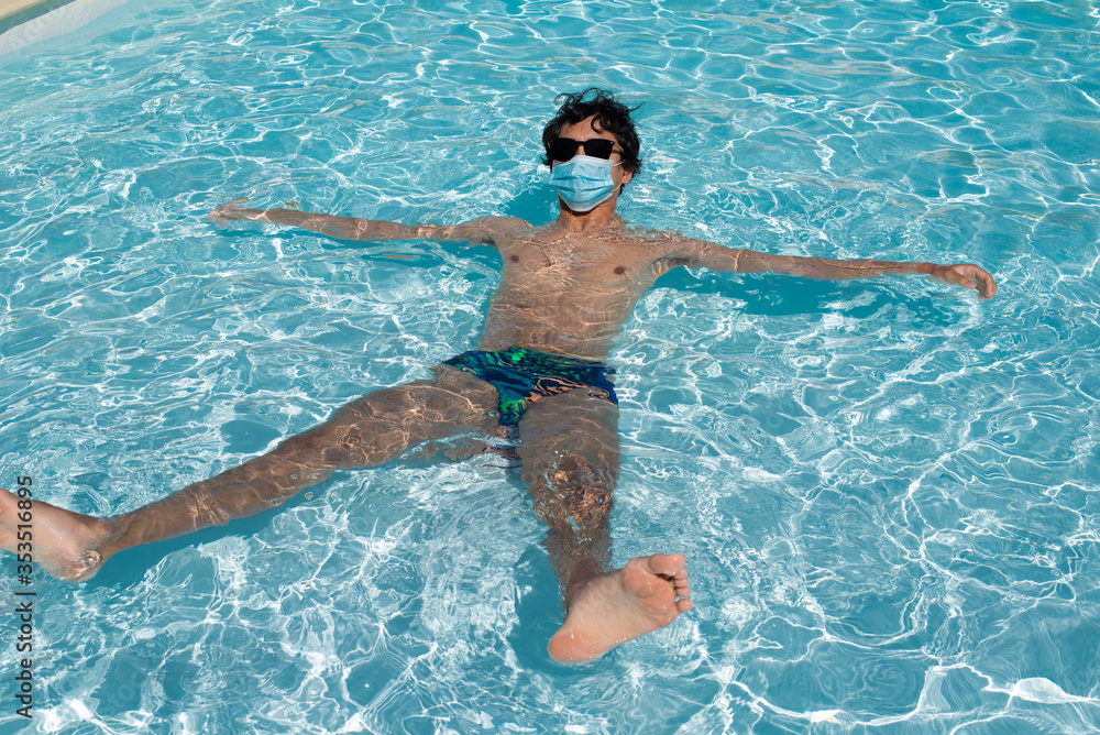Young handsome man floating in a swimming pool wearing a protective mask during coronavirus summer