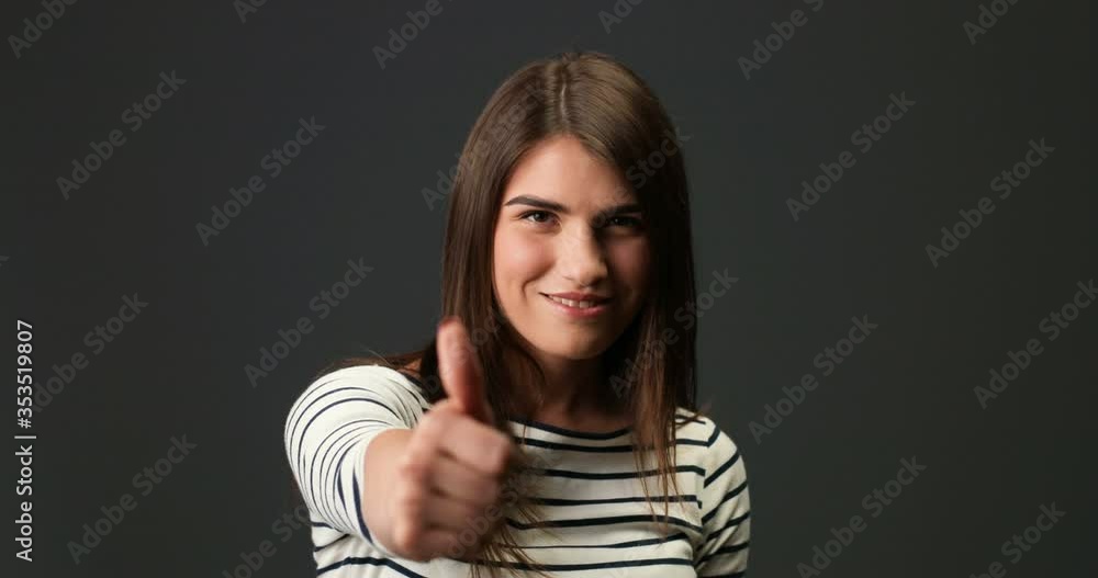 Close up portrait of cheerful beautiful Caucasian young woman in striped blouse with smile on face standing and giving thumb up. Happy pretty girl with dark hair on grey background. Emotion concept Close up portrait of cheerful beautiful Caucasian young woman in striped blouse with smile on face standing and giving thumb up. Happy pretty girl with dark hair on grey background. Emotion concept