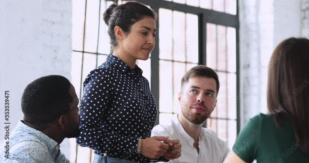 Happy young indian businesswoman standing at table, sharing project ideas with focused mixed race investors at briefing meeting. Professional hindu female mentor coaching millennial colleagues.