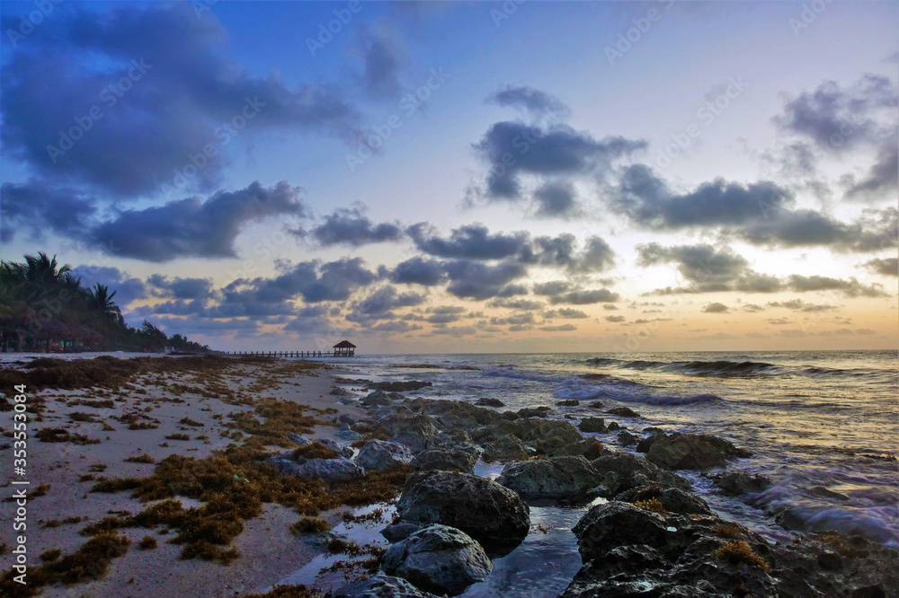 Beautiful sunrise in mexico. The clouds are backlit by the sun in shades of pink and orange. At sea, the waves. In the sand, large stones and algae. In the distance is a wooden platform and a canopy.