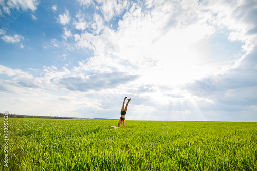 Girl acrobat performs a handstand. The model stands on her hands, doing gymnastic splits against the background of green trap and blue sky, playing sports in nature