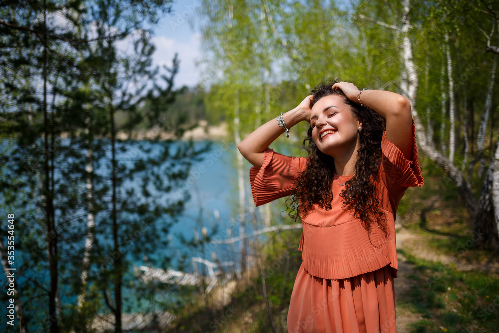 Naklejka premium A girl with long wavy curly hair in her hair woven flowers in a white transparent guipure dress barefoot in the summer on nature in a forest at sunset standing standing near the road and bushes
