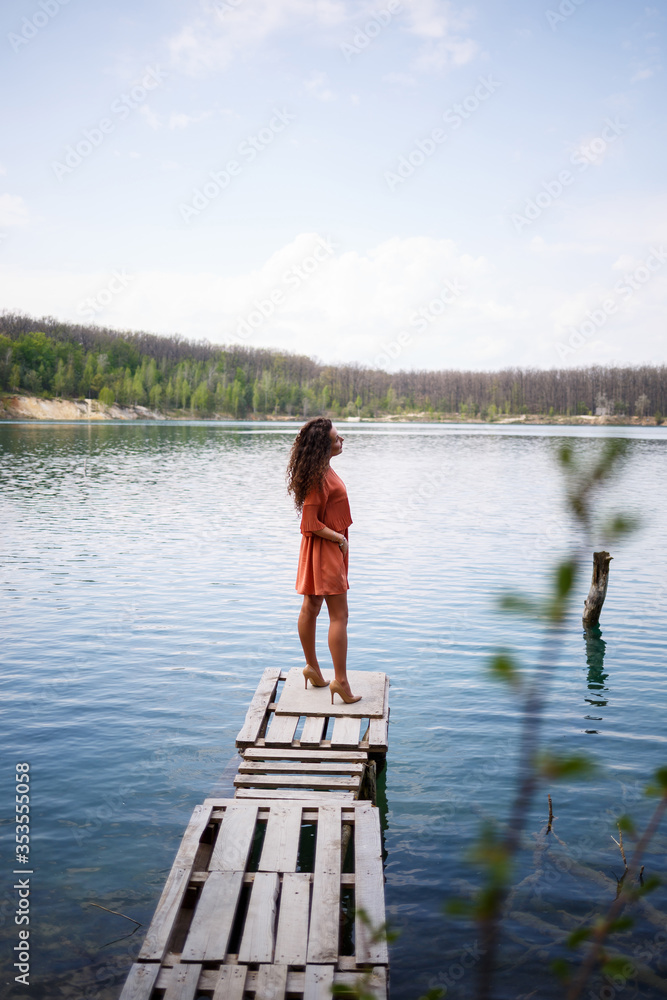 Wooden masonry. A girl with long wavy curly hair in an orange guipure dress and shoes on nature, in a forest against a lake, stood near trees and bushes. Young woman smiles and enjoys life