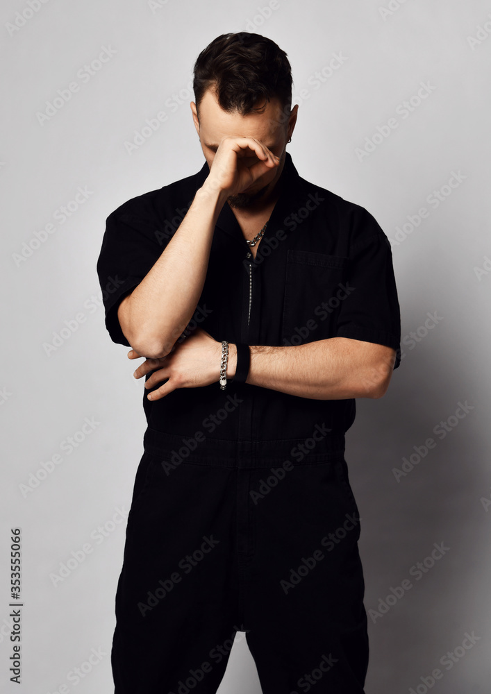 Portrait of young brutal man in black casual clothing denim overall stands looking down with hand at his elbow Portrait of young brutal man in black casual clothing denim overall stands looking down with hand at his elbow