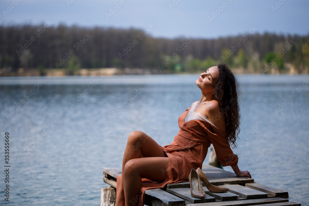 A girl with long wavy curly hair in a long guipure dress barefoot in the summer in a forest on a lake at sunset standing on a pantone on a wooden pier bridge. Summer sunny day