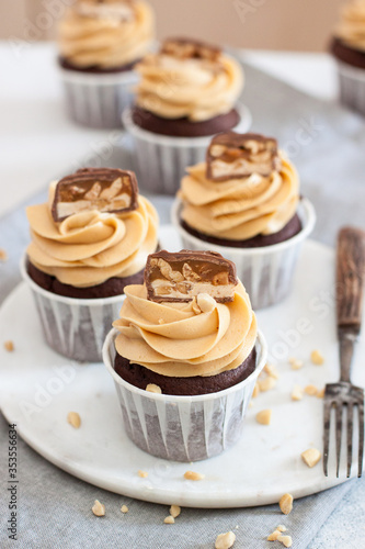 Peanut butter cupcakes with chocolate bites and chopped nuts on grey linen tablecloth.