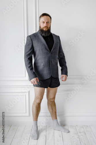 bearded man in a jacket and without pants stands against a light wall