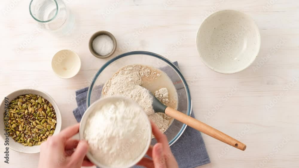 Woman hands adds flour in yeast water, kneading dough for whole grain bread from whole wheat flour, flax, sunflower and pumpkin seeds close-up on a wooden background. Top view. Wholemeal Bread. Woman hands adds flour in yeast water, kneading dough for whole grain bread from whole wheat flour, flax, sunflower and pumpkin seeds close-up on a wooden background. Top view. Wholemeal Bread.