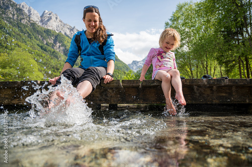 Mother and daughter sitting on wooden pier having fun splashing water with their feet.