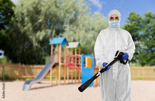 disinfection service and pandemic concept - sanitation worker in protective gear, medical mask, gloves and goggles with pressure washer or sanitizer over children's playground background